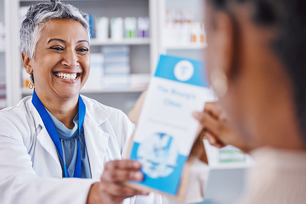 A woman in a white coat is handing an item to a customer at a pharmacy counter.